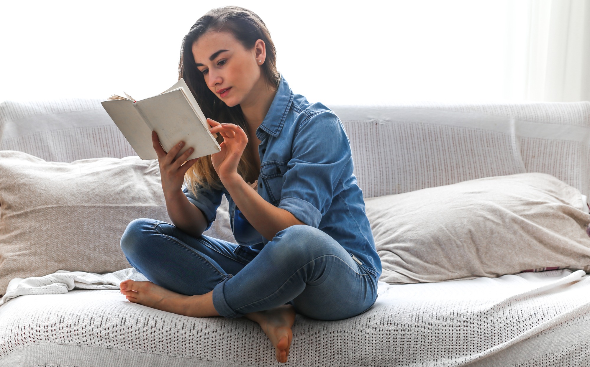 A woman sits on a couch, engrossed in reading a book, surrounded by a cozy living room atmosphere.